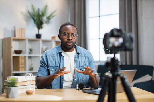 African American Man In Denim Shirt And Eyeglasses Sitting At Table, Gesturing And Talking While Recording Video On Camera. Male Blogger Doing Live Stream In His Social Network.