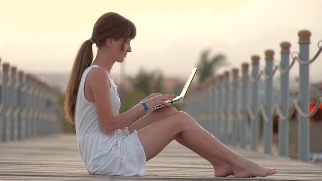 Young female office worker working on laptop computer outdoors on warm summer evening. Remote work and study concept