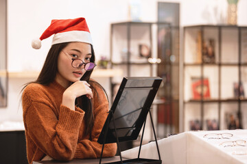 Beautiful girl wearing Santa hat is choosing a glasses in optician store, Asian girl in sweater...
