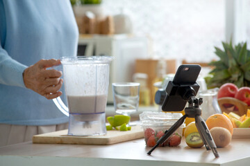 Close up hands of senior woman blogger prepares a healthy drinks in front of smart phone on tripod, older women and technology concept