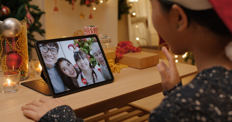 Asia happy woman relax smile laugh waving hand greet in digital tablet screen remote talk to child friend at night online VoIP X'mas party sit on cozy sofa at home enjoy good warm time winter season.