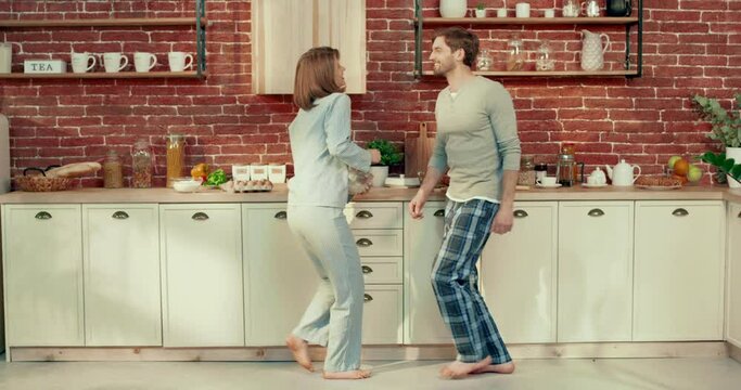 Happy man and woman dancing in kitchen in pajamas, moving synchronously while preparing breakfast. Harmony of married couple.