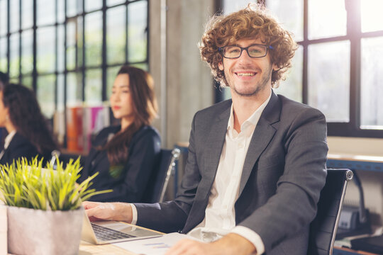 Portrait Of A Happy Caucasian Male Employee Posing For A Corporate Business Catalog With Coworkers In The Backdrop During A Photo Shoot In The Office.