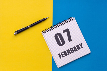 February 7th. Day 7 of month. Calendar date. Notebook with a spiral and pen lies on a yellow-blue background