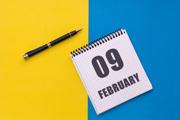 February 9th. Day 9 of month. Calendar date. Notebook with a spiral and pen lies on a yellow-blue...
