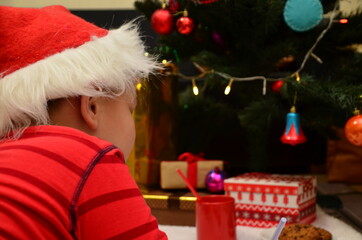 Christmas Child Write Letter to Santa Letter. Kid in Xmas Hat Writing Gift Wish List next to Christmas Tree over dark Background