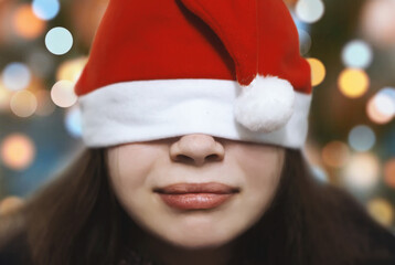 Close-up of a happy young woman in a red Santa hat pulled over her eyes, smiling sweetly. A young...