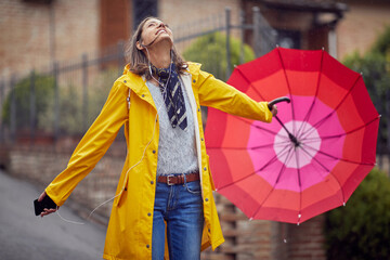 A young cheerful woman in a yellow raincoat is looking to the sky while listening to the music and walking the city on a rainy day. Walk, rain, city