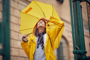 A young cheerful woman with a yellow raincoat and umbrella who is enjoying while listening to the music and walking the city on a rainy day. Walk, rain, city - obrazy, fototapety, plakaty