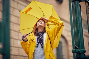 Fotobehang A young cheerful woman with a yellow raincoat and umbrella who is enjoying while listening to the music and walking the city on a rainy day. Walk, rain, city © luckybusiness
