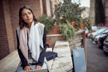 A young elegant and attractive woman is thoughtfully sitting in the street and using laptop. Walk, outside, rest
