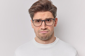 Obraz premium Sulking dissatisfied adult European man looks sadly at camera frowns angrily wears transparent glasses and casual jumper isolated over white background stands upset complains about something