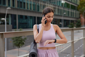 Sporty young woman checks time on smarwatch makes telephone call via smartphone dressed in sportswear carried karemat stands outside poses outdoors against blurred background waits for coach