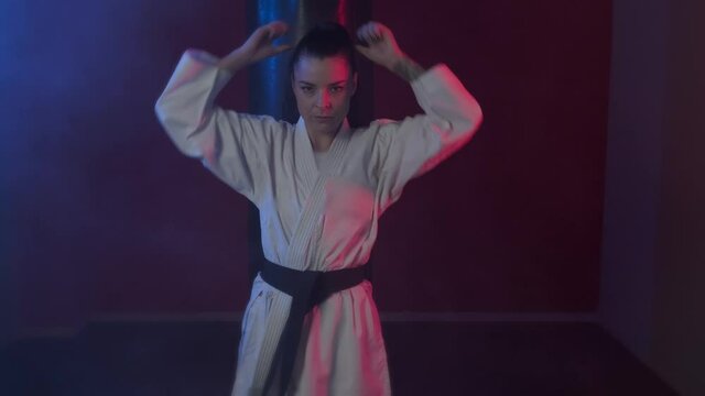 A young karate girl in a kimono straightens her hair, tightens her belt before the fight and looks seriously at the camera. Strong woman practicing karate, is ready for a fight in a dark sports hall. 