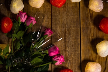 Romantic surprise for celebrating Valentine's Day made of roses and garland with wine glasses. Preparing for engagement event. Nice pink flowers and flashing light with heart shape lie on wooden table