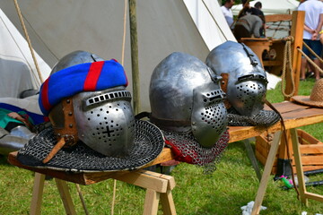 A close up on three well maintained medieval helmets with neck protection chainmail laying on a wooden table next to a cloth tent seen during a historic fair in Poland on a sunny summer day