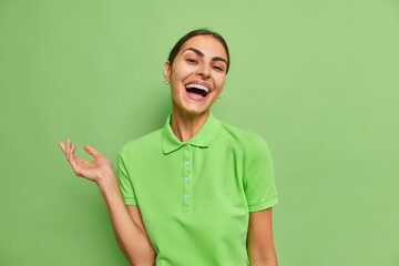 Waist up shot of happy brunette woman dressed in casual t shirt smiles sincerely keeps arm raised...