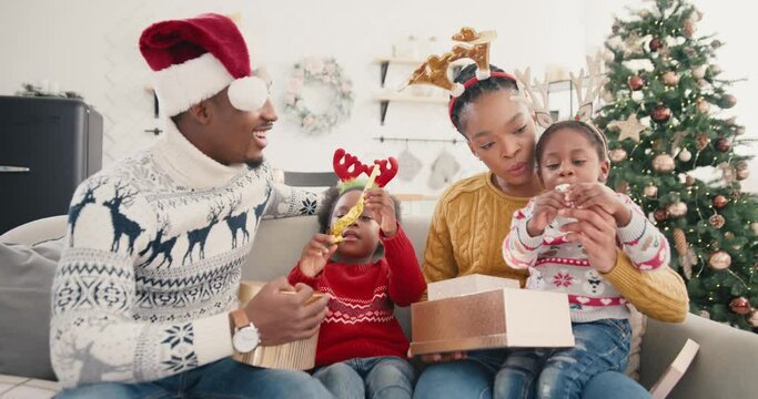 Portrait of curious African American kids open gift boxes while sitting with happy parents together at decorated home near pretty Christmas tree New Year Eve. Holidays celebration xmas present concept
