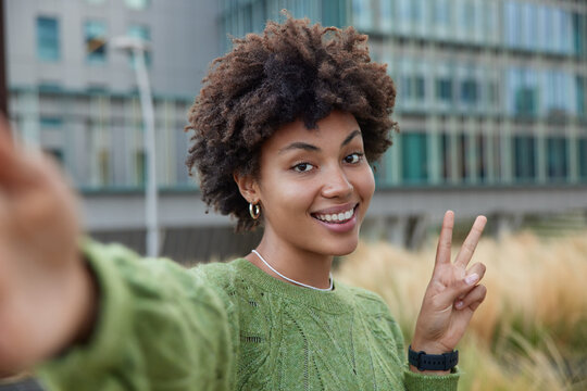 Happy young woman enjoys spare time makes peace sign stretches arm makes selfie smiles gladfully wears casual green jumper enjoys good day poses outdoors against blurred background shoots video