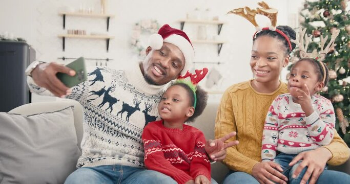 Portrait of happy African American family taking pictures on smartphone and smiling in christmassy decorated home. Dad in Santa hat taking selfie photo on cellphone with kids and wife