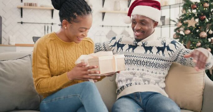 Portrait of happy loving husband making surprise to his wife on Christmas Eve and giving xmas present. Joyful wife getting gift box from caring beloved man near xmas tree at home. 2022 New Year