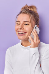 Vertical shot of happy young woman talks via mobile phone with best friend smiles gladfully enjoys nice conversation dressed in casual white jumper isolated over purple background. Communication