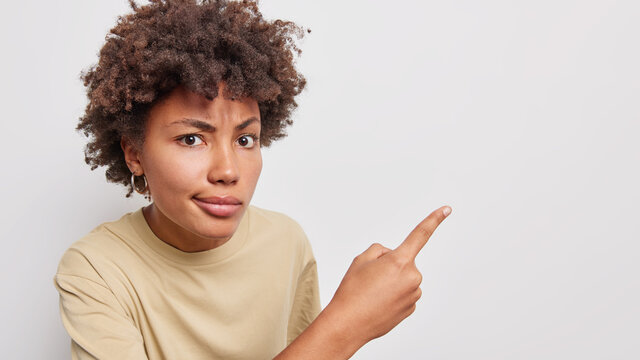 Serious curly haired woman shows something unpleasant points with finger at blank wall shows product advertisement recommends to try item subscribe or click on link isolated over white background