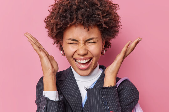 Positive emotional young pretty woman with curly hair raises palms near gead exclaims loudly keeps eyes closed wears black striped jacket reacts on wonderful news isolated over pink background.