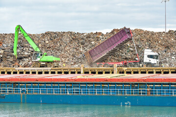 Machine operated grabbing arm, loading scrap metal onto a ship in the harbour of Newhaven, UK, with lorry delivering more scrap metal to the dock yard.