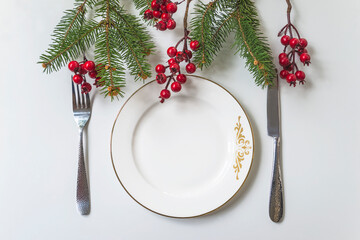 Festive place setting for christmas dinner; Empty white plate, fork, knife and fir tree twigs, red berries on white background top view