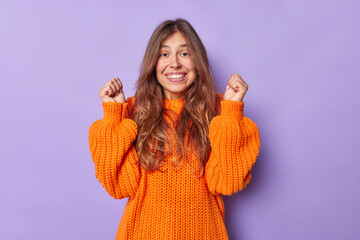 Happy excited woman rejoices success clenchs fists and smiles gladfully wears casual knitted...
