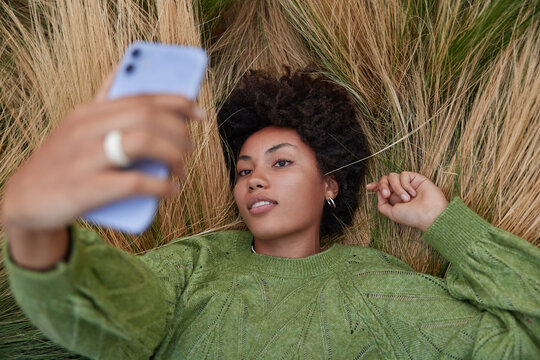 Overhead Shot Of Relaxed Curly Haired Young Woman Dressed In Casual Jumper Lies Relxed In Long Grass Holds Mobile Phone Makes Selfie Portrait Spends Free Time Outdoors. Millennial Content Maker