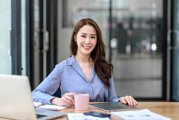 Beautiful young Asian businesswoman holding a coffee mug working at office. Looking at camera.