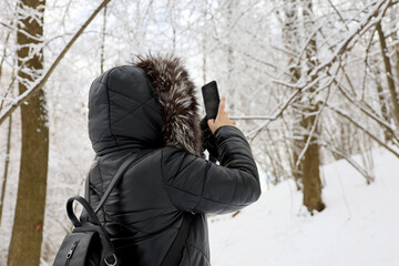Fototapeta na wymiar Woman in black coat with fur hood taking pictures of snow nature on a smartphone in the winter forest. Trees after snowfall, leisure at cold weather