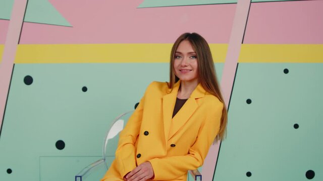 Bright stylish woman, blue-eyed European with charming smile in yellow suit is sitting in transparent chair in creative space, looking at camera and sharing positive vibes, Zoom in, Slow motion.