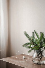 Spruce branches and a plant with white round balls on twigs in a large glass vase on a wooden surface against a wall and transparent tulle, surrounded by a Christmas garland.