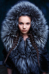 Beautiful fashionable girl in a fur coat with a hood is posing in the studio. Closeup portrait of a stylish kid, dark background, twilight  - 472984756