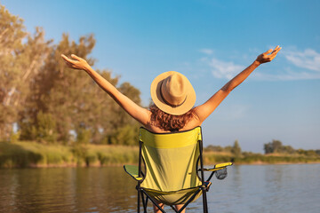 Happy woman with hat sitting on a camping chair on the shore of a river or lake. Outdoor recreation and relaxation