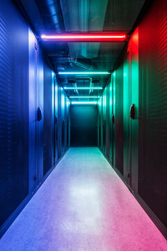 Cold aisle of an IT server room with perforated grille doors illuminated in color and large cooling pipes on the ceiling