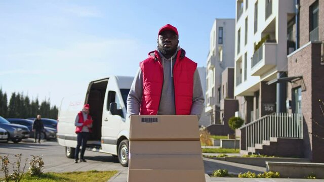 African american delivery man carring parcel boxes on a trolley from a van. Courier on the way to deliver postal parcel to a client.