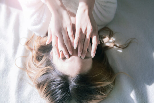 白い明るい部屋のベッドで朝日を浴びながら目の疲労をマッサージする女性