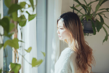 Fototapeta premium 植物と光の差し込む窓辺に佇み、外を眺めるロングヘアの美しい女性