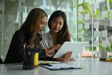 Photo of two female designers working together with a digital tablet at the white working desk in the comfortable living room.