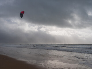 A kitesurfer surfing along the waves on a sandy beach against a cloudy and stormy sky in Wales