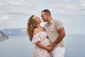 happy and very beautiful young couple expecting a baby. a pregnant woman laughs, and a happy man holds her belly against the backdrop of a beautiful seascape