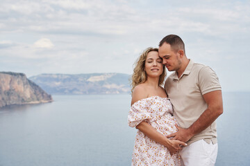 Fototapeta premium beautiful couple on the background of a beautiful seascape. a man gently holds a pregnant woman by the stomach. smiling woman