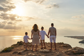 a large family is holding hands against the backdrop of a sunset sky and a seascape with a rocky coastline stretching into the distance. back view