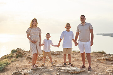 young family on the seashore in warm rays. pregnant mom, dad and two sons hold hands
