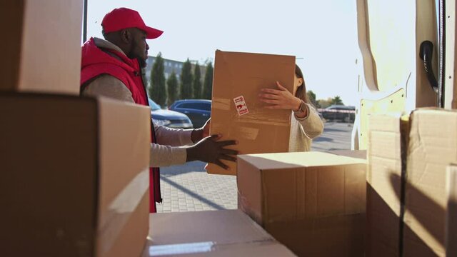 African american courier in red uniform taking parcel box from a van and give it to a client. Caucasian woman take an order and signed on a tablet.
