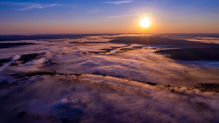 Fototapeta premium Aerial view of a beautiful summer landscape with a fog while dawn. Photo from drone of a foggy landscape in spring. Top view to land while sunrise. Sun is above the horizon